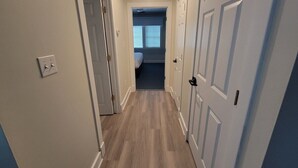 A narrow hallway with light-colored walls and wooden flooring, leading to a room with a bed and a window with blinds. - The family -friendly resort life is waiting for you! 
 (Williamsburg)