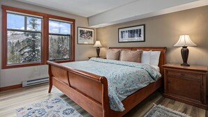 Een slaapkamer met een houten bed, twee bedlampen, een nachtkastje en een groot raam met uitzicht op bomen.