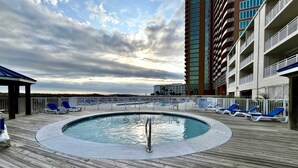 Zona de piscina con muelle de madera, sillas de descanso y un edificio alto al fondo.