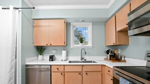 Una cocina moderna con armarios de madera clara, lavavajillas de acero inoxidable, horno integrado y ventana con vista a palmeras.