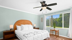 Un dormitorio con una cama de madera, dos mesitas de noche con lámparas, un ventilador de techo, una ventana con cortinas y una pequeña mesa de madera con una planta.