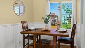 Un comedor con una mesa de madera, dos sillas, un jarrón y una ventana que da a una casa y palmeras.