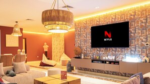 Un soggiorno moderno con una grande TV a schermo piatto che mostra il logo di Netflix, un'area salotto accogliente con divani e un tavolino, e una parete decorativa con un motivo geometrico.