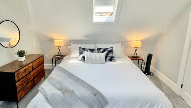 Un dormitorio con una cama grande, dos lámparas de noche, una lámpara de techo, un tragaluz, un aparador de madera y un espejo en la pared.