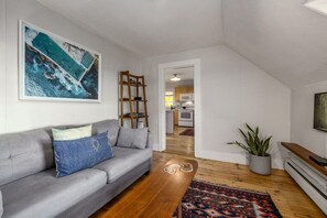 A modern living room with a grey sofa, wooden coffee table, and a colorful rug. A large painting is on the wall, and a plant is in a grey pot.