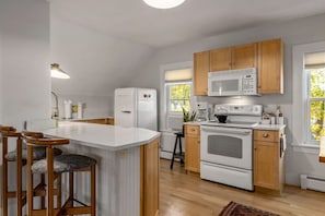 A kitchen with white appliances, wooden cabinets, a central island with stools, and a view through the window.