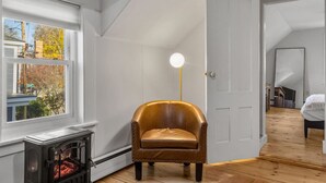 Une pièce avec une cheminée, un fauteuil en cuir, un lampadaire rond et un lit recouvert d’une couverture blanche.