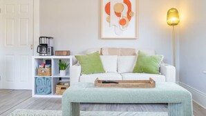 Ein Wohnzimmer mit einem weißen Sofa, grünen Kissen, einer Kaffeemaschine, einem gerahmten abstrakten Kunstwerk und einer Stehlampe.