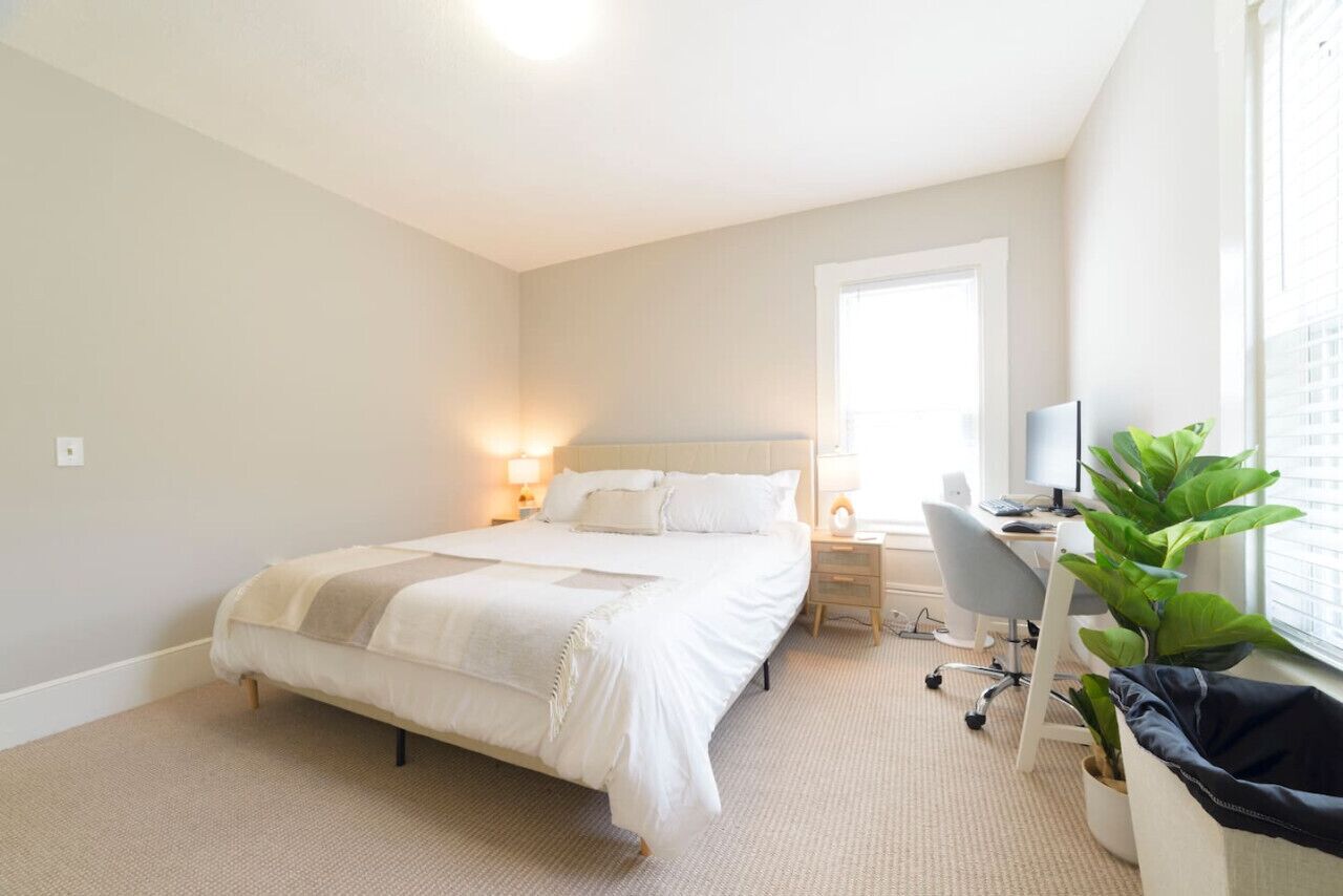 Une chambre d’hôtel avec un grand lit, un bureau équipé d’un ordinateur, une chaise et une fenêtre avec des stores.
