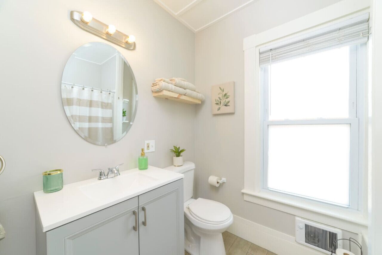 A bathroom with a round mirror, a white sink, a toilet, and a window.