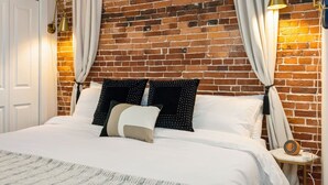 A bedroom with a brick wall, white bedding, and two bedside lamps. - Tugboat Vista 2 Bedroom Downtown Portsmouth (Portsmouth)