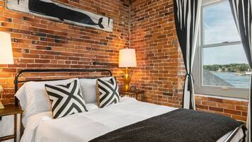 A room with a brick wall, a bed with white bedding and patterned pillows, a nightstand with a lamp, and a window with curtains.