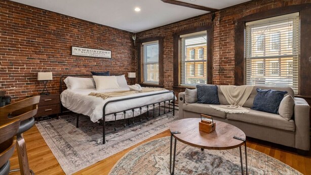 Una habitación con pared de ladrillo, una cama, un sofá, una mesita de café y una ventana con persianas.
