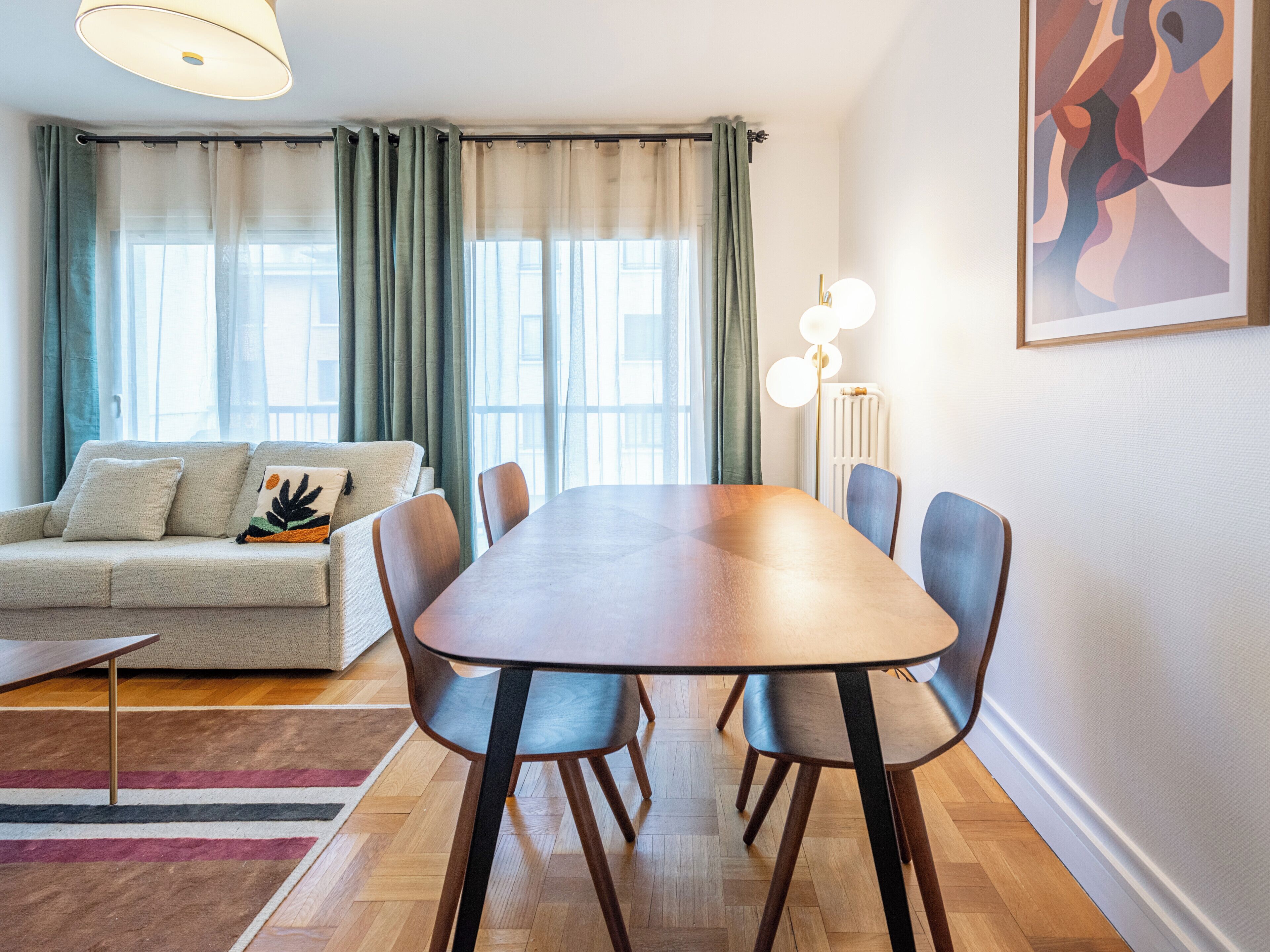 A modern living room with a sofa, a dining table, and chairs. There is a window with curtains and a radiator.