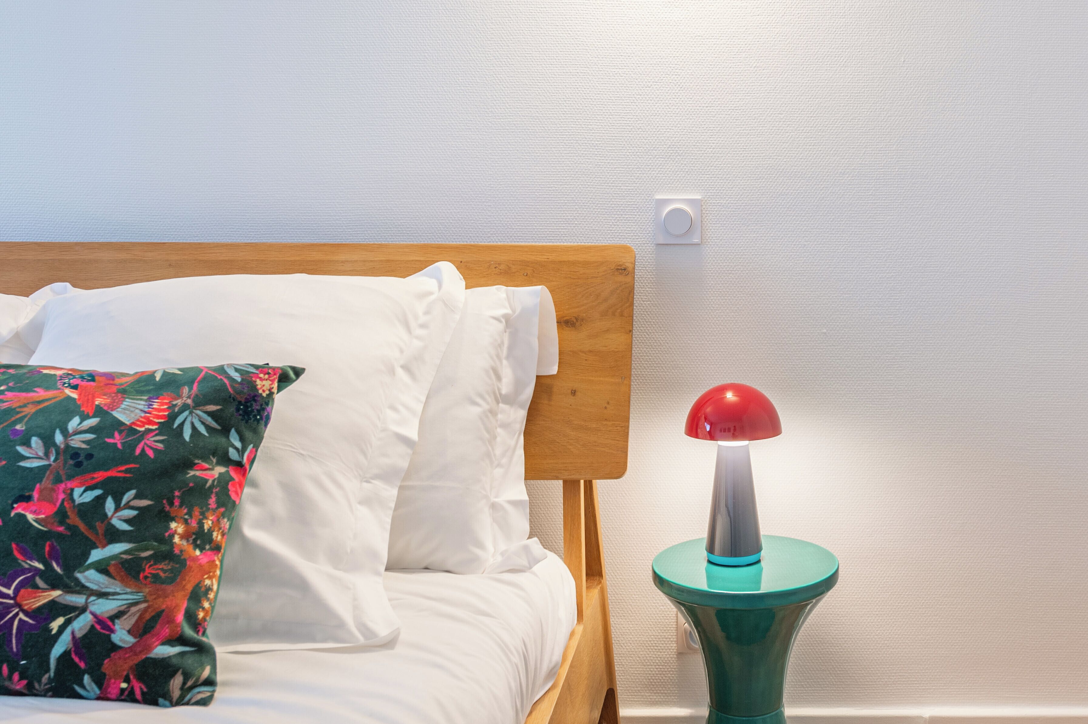A bed with white bedding and a colorful floral pillow, a wooden headboard, and a small teal side table with a red lamp.