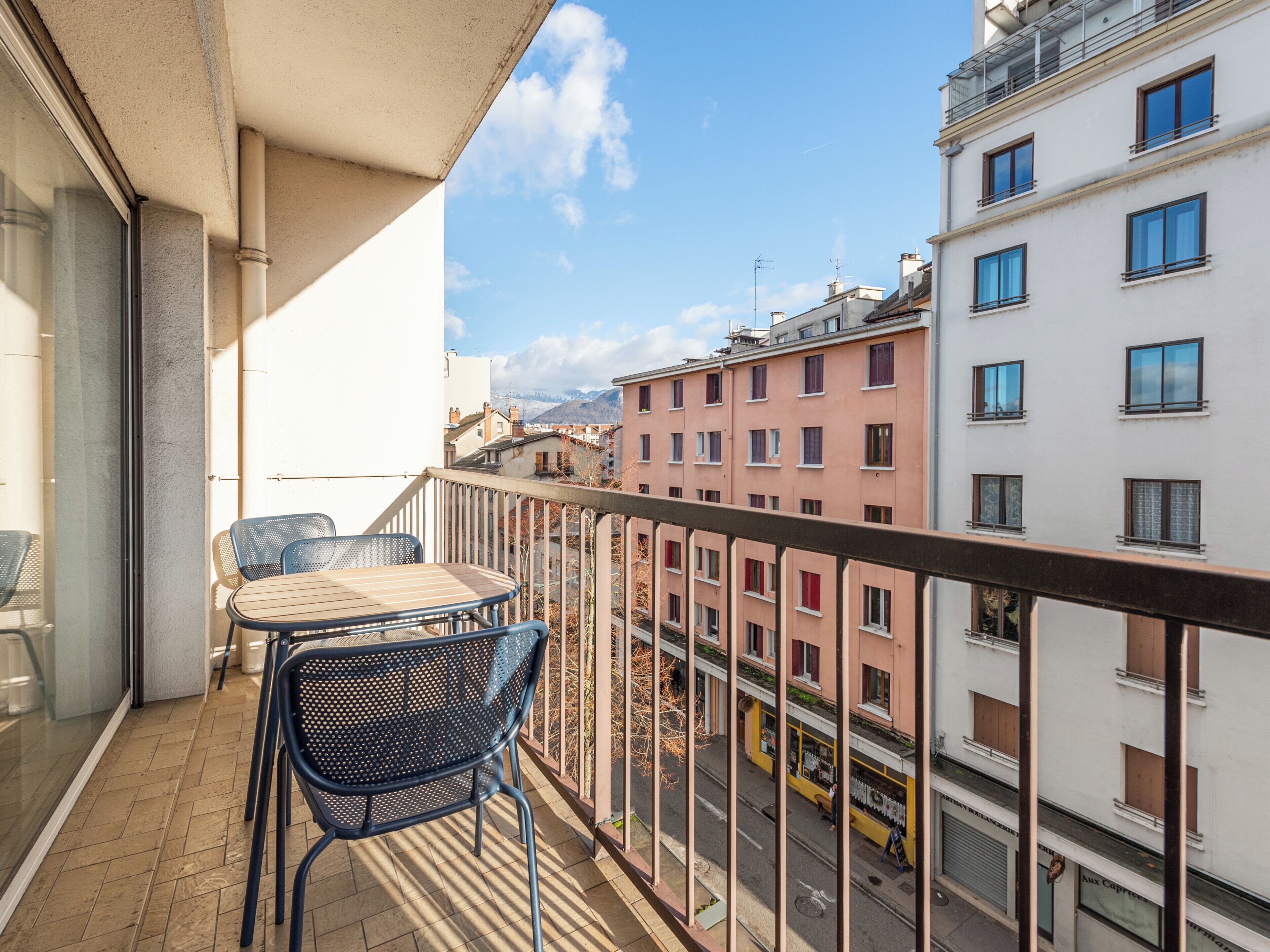 Un balcón con mesa y sillas, con vista a una calle con edificios y cielo despejado.