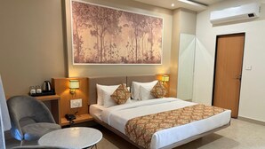 A hotel room with a bed, bedside tables, a chair, a small table, a painting on the wall, and an air conditioning unit. - Hotel Aura By Arihant (Udaipur)