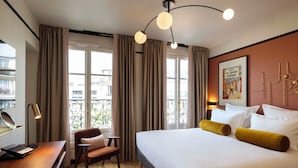 A hotel room with a large bed, a desk with a laptop, a chair, and a view of the city through large windows. - Les Rives Oceanik (Paris)