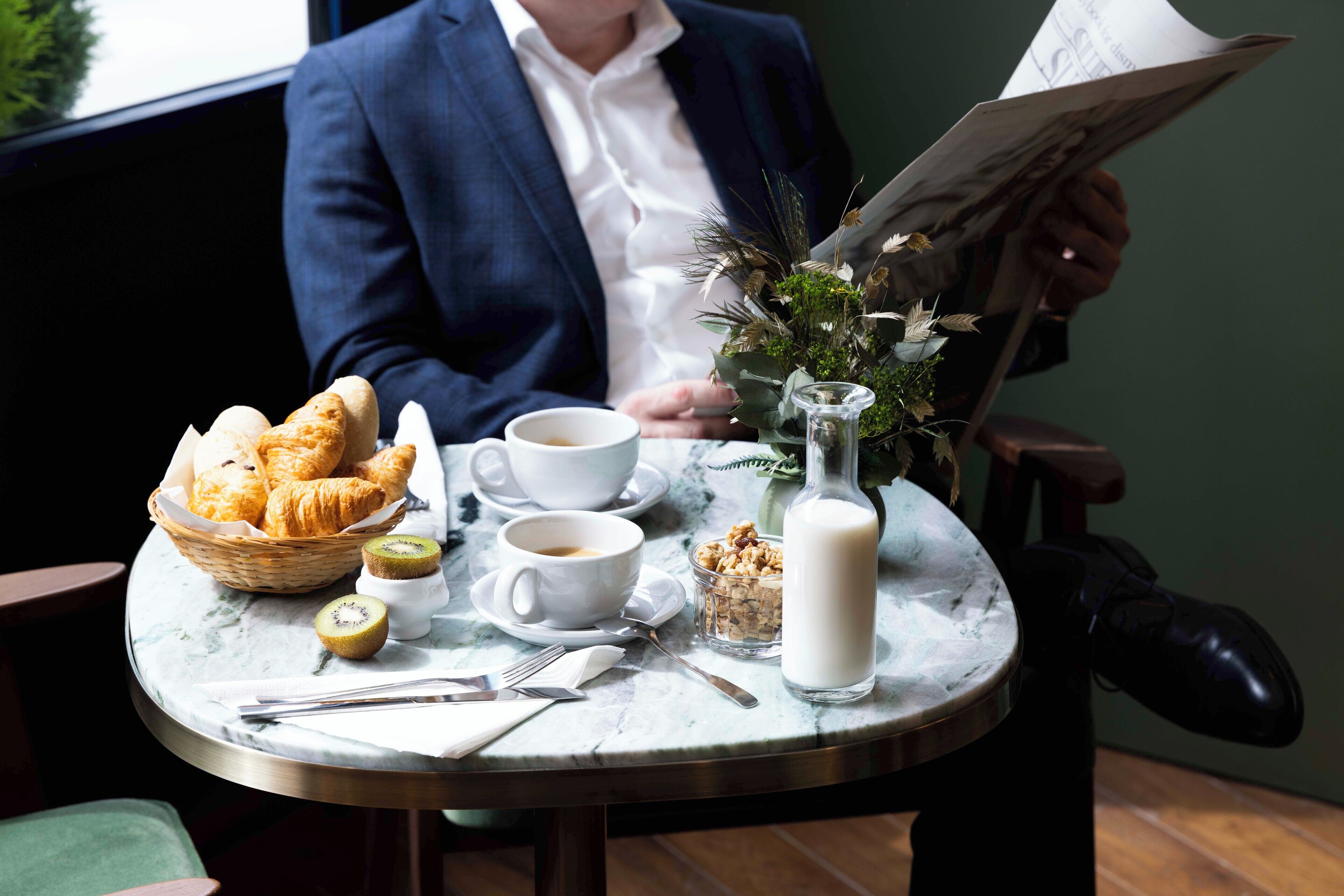 A breakfast setting with croissants, coffee, milk, and a newspaper on a marble table.