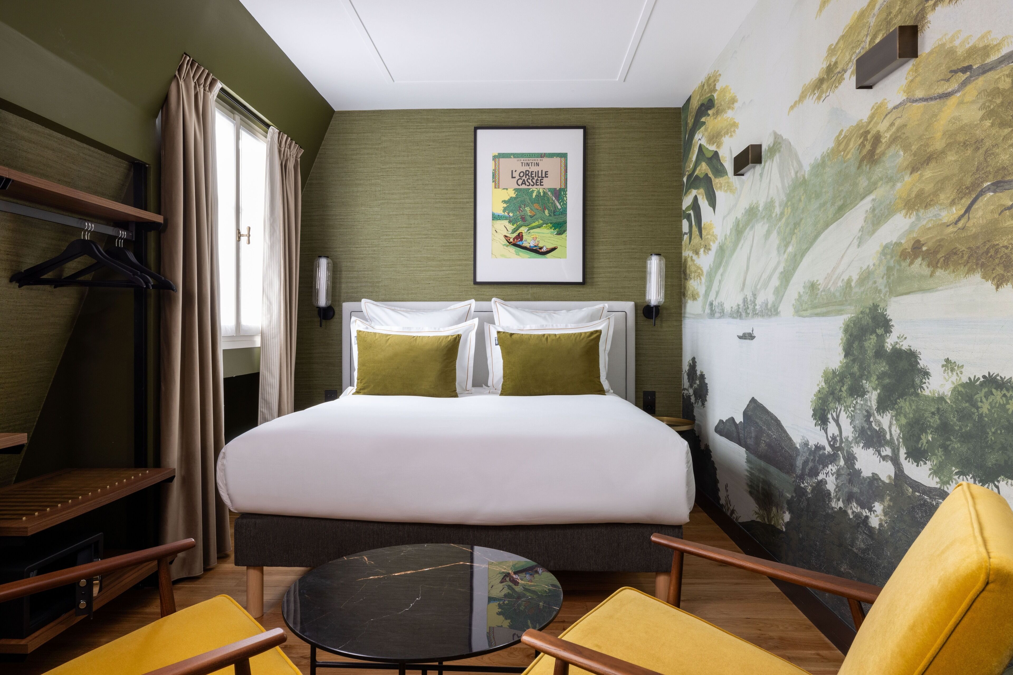 Een hotelkamer met een bed, twee gele stoelen, een klein tafeltje, een schilderij aan de muur en een raam met gordijnen.
