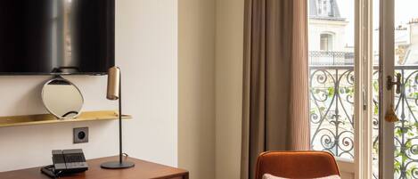 A hotel room with a wooden desk, a chair, a television, and a view of the city through a balcony door.