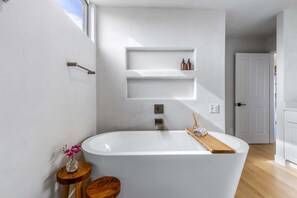 A modern bathroom with a freestanding bathtub, wooden side table, and a small shelf with toiletries.