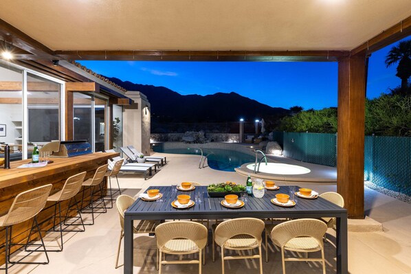 An outdoor dining area with a pool, chairs, and a table set for a meal.