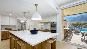 Una cocina moderna con una isla central, taburetes de madera y vistas a una piscina y montañas.