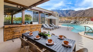 Un área de comedor junto a la piscina con una mesa para cuatro, bar al aire libre, sillas de descanso y vistas a las montañas.
