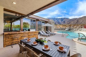 A poolside dining area with a table set for four, outdoor bar, lounge chairs, and a view of mountains.