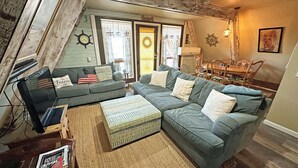 Una sala de estar acogedora con un sofá modular, una mesita de centro y una alfombra. En el fondo, se observa un comedor con sillas.