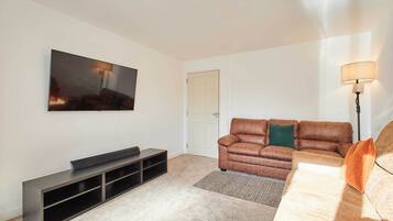 A living room with a flat-screen TV, a brown leather sofa, a wooden coffee table, and a floor lamp.