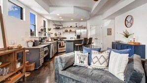 A modern kitchen with a built-in oven, a dining area with a grey sofa, and a wall-mounted clock.