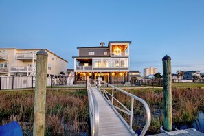 Een twee verdiepingen tellend strandhuis met een terras dat naar de ingang leidt, omringd door water en begroeiing.