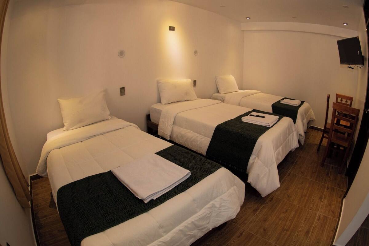 A hotel room with three single beds, each with white linens and a dark green throw blanket. There is a wooden chair and a flat-screen TV mounted on the wall.