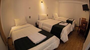 A hotel room with three single beds, each with white linens and a dark green throw blanket. There is a wooden chair and a flat-screen TV mounted on the wall.