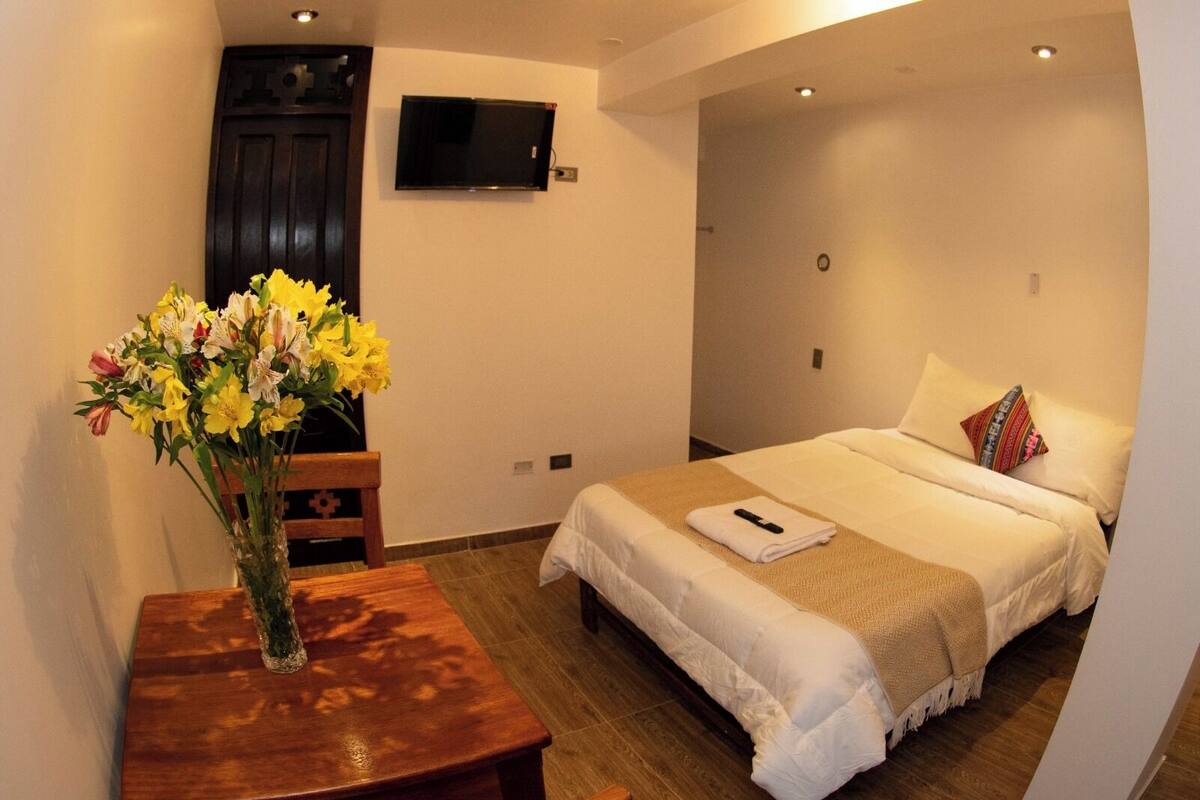 A hotel room with a bed, a TV mounted on the wall, a wooden table with a vase of flowers, and a small desk.