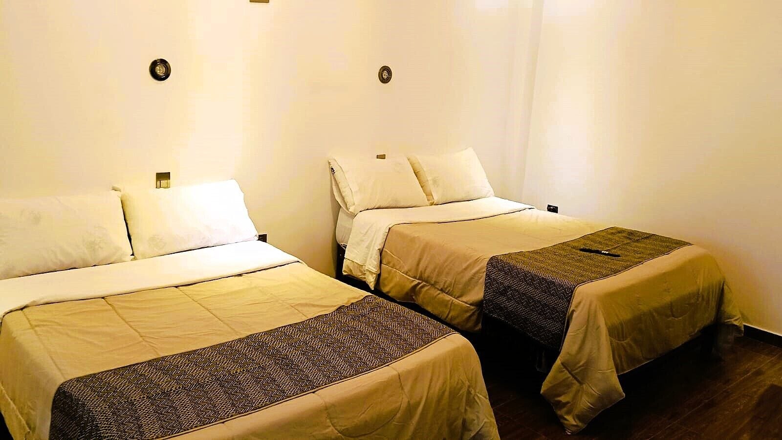 Dos camas individuales con ropa de cama color crema y almohadas decorativas estampadas, una mesita de noche pequeña y un aplique de pared.