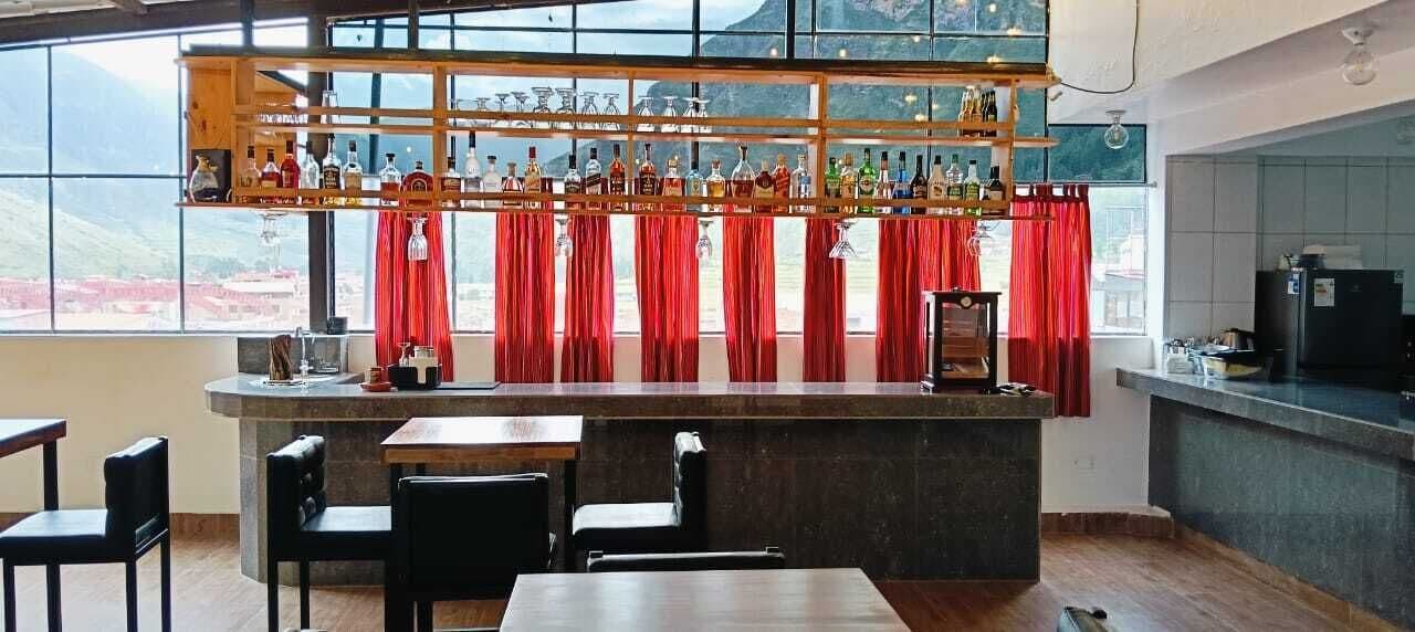Un espace bar avec un long comptoir, de nombreuses bouteilles et des rideaux rouges. On y trouve des tabourets de bar et une table avec des chaises.