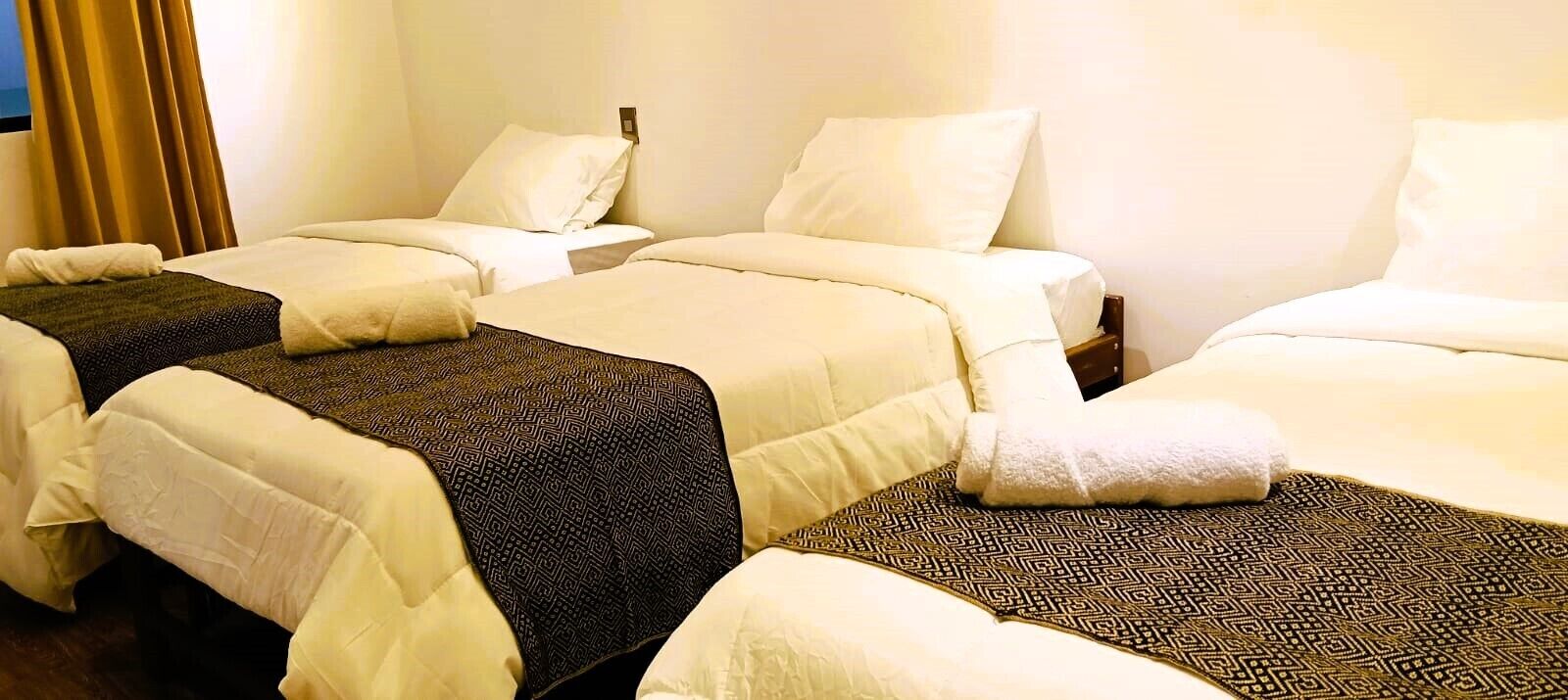 Habitación de hotel con tres camas dispuestas en fila, cada una con ropa blanca y una manta estampada.