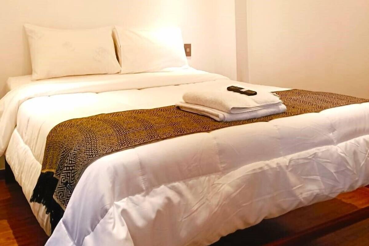 A hotel room with a bed, a flat-screen TV mounted on the wall, and a bedside table with a lamp.