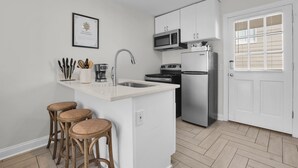 A modern kitchen with white cabinetry, a stainless steel refrigerator, a microwave, and a sink. There are three wooden stools and a framed certificate on the wall.