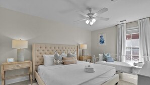A bedroom with a large bed, two bedside tables, a ceiling fan, a window with curtains, and a wall-mounted artwork.