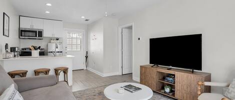 A modern living room with a sofa, a coffee table, a TV on a wooden cabinet, and a kitchen area in the background.