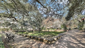 A paved pathway with benches and a grassy area, surrounded by trees and buildings.