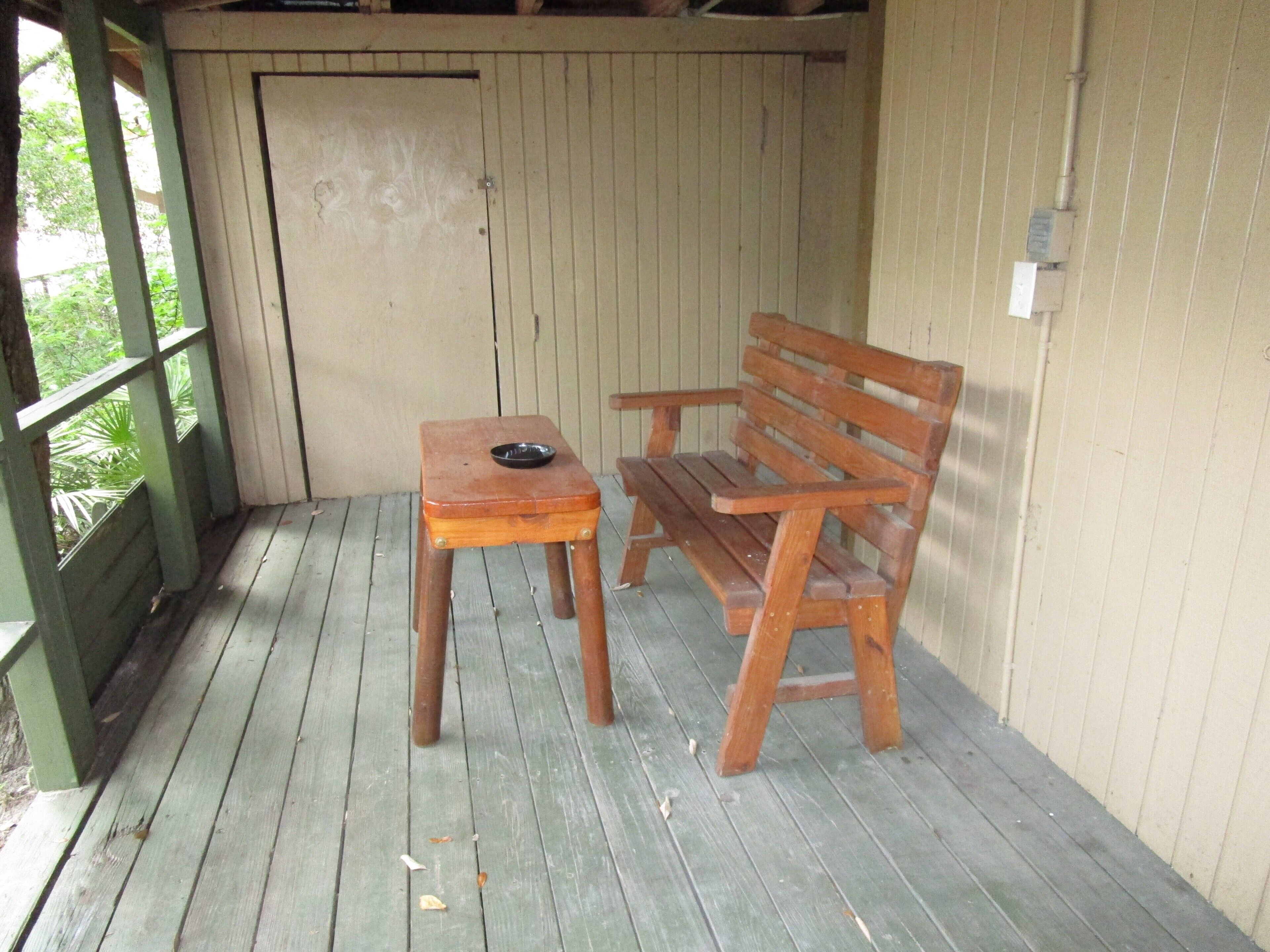 A wooden bench and table on a wooden deck with a green railing, a beige wall, and a door in the background.
