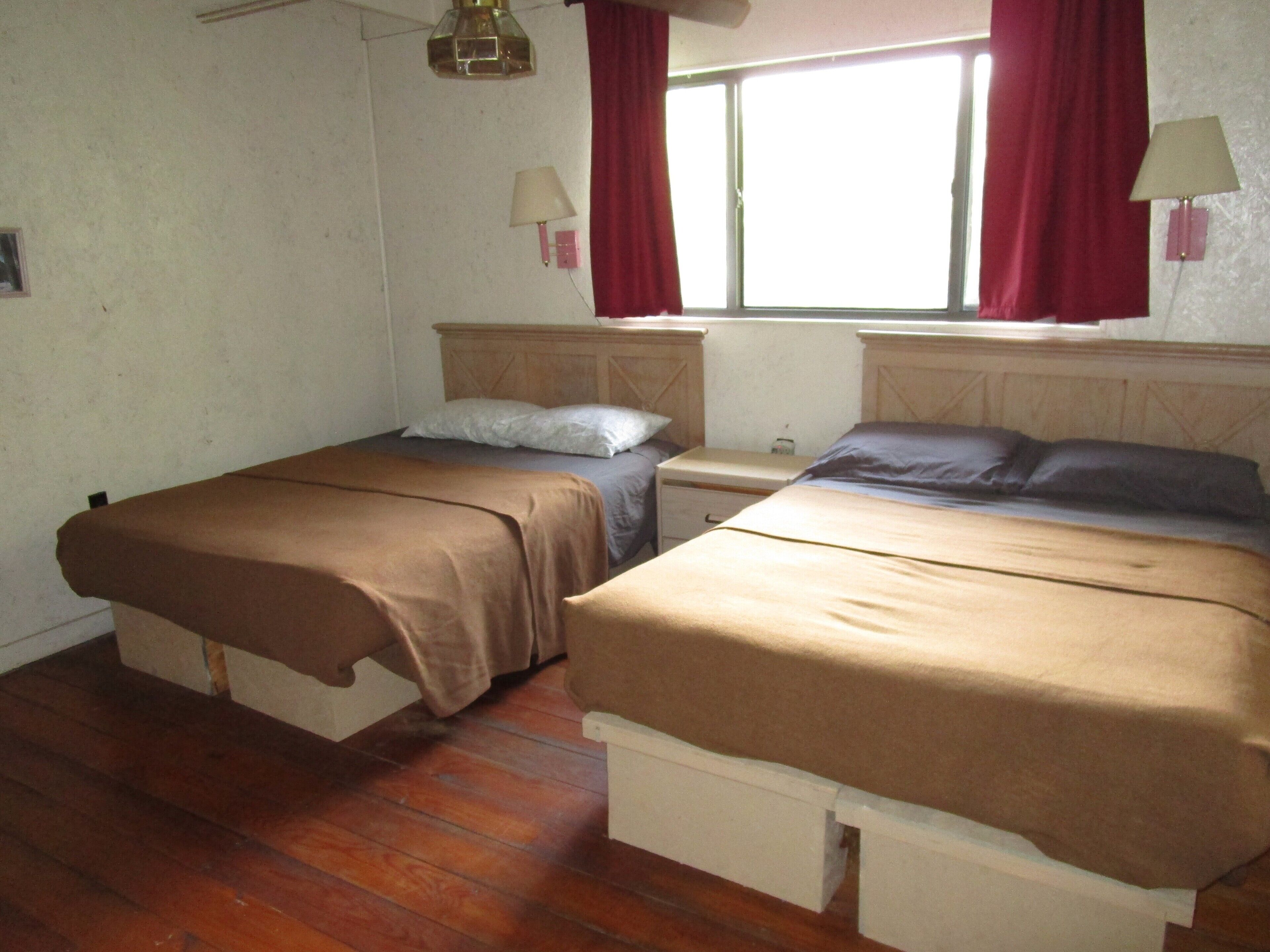 A room with two beds, wooden flooring, a window with red curtains, and a wall-mounted lamp.