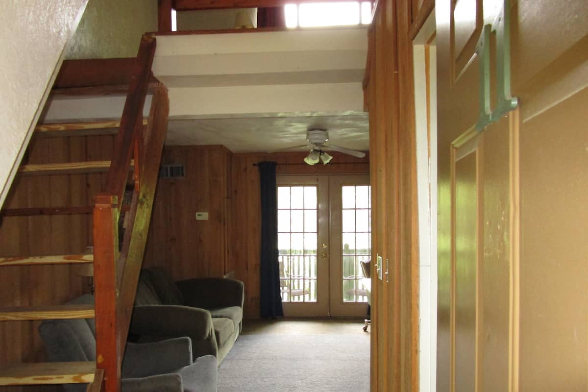 A staircase leading to a second floor, a door with a window, and a ceiling fan in a room with wooden walls and carpeted floor.