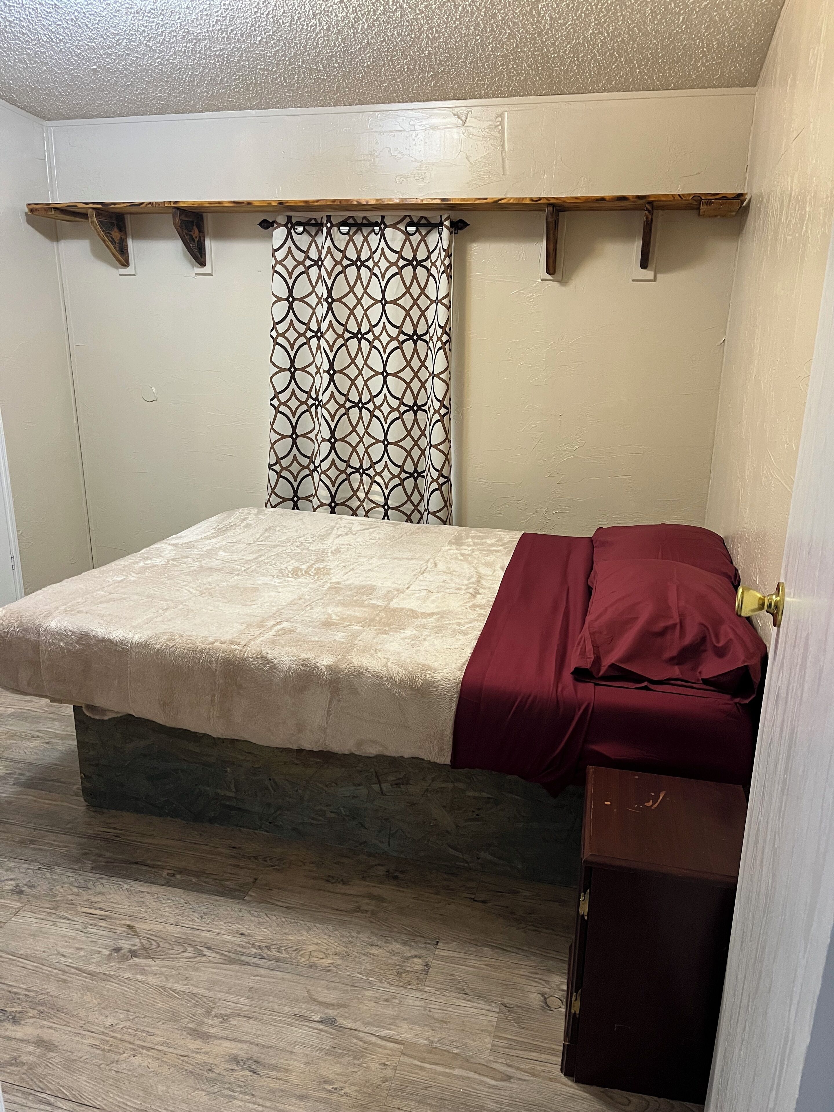 Een slaapkamer met een bed, een houten bankje, een gordijn met patroon en een klein kastje.