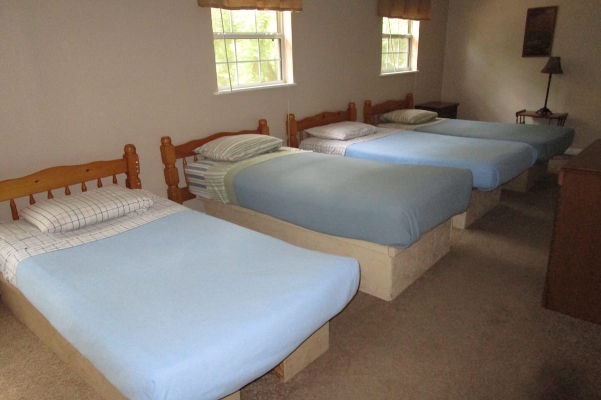 A room with three single beds, each with a wooden headboard and blue bedding. There are two windows with curtains, and a lamp on a bedside table.
